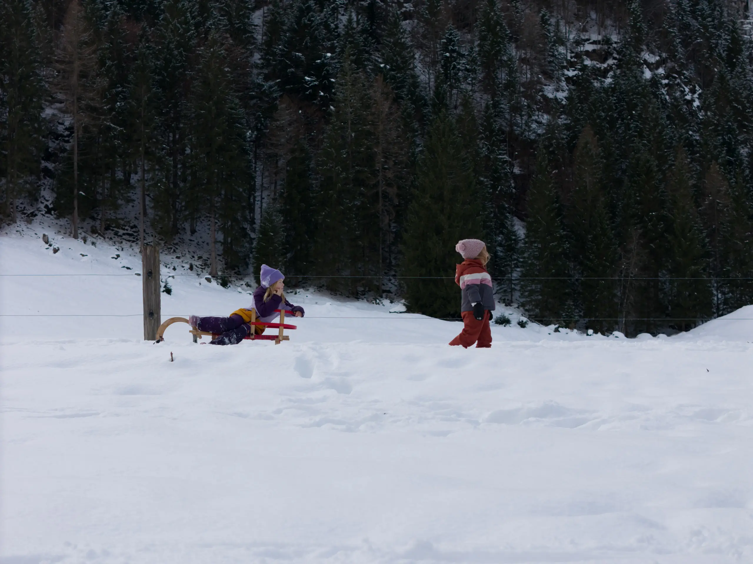 Zwei Kinder spielen im Schnee, eines auf einem Schlitten vor einem bewaldeten Berghang.