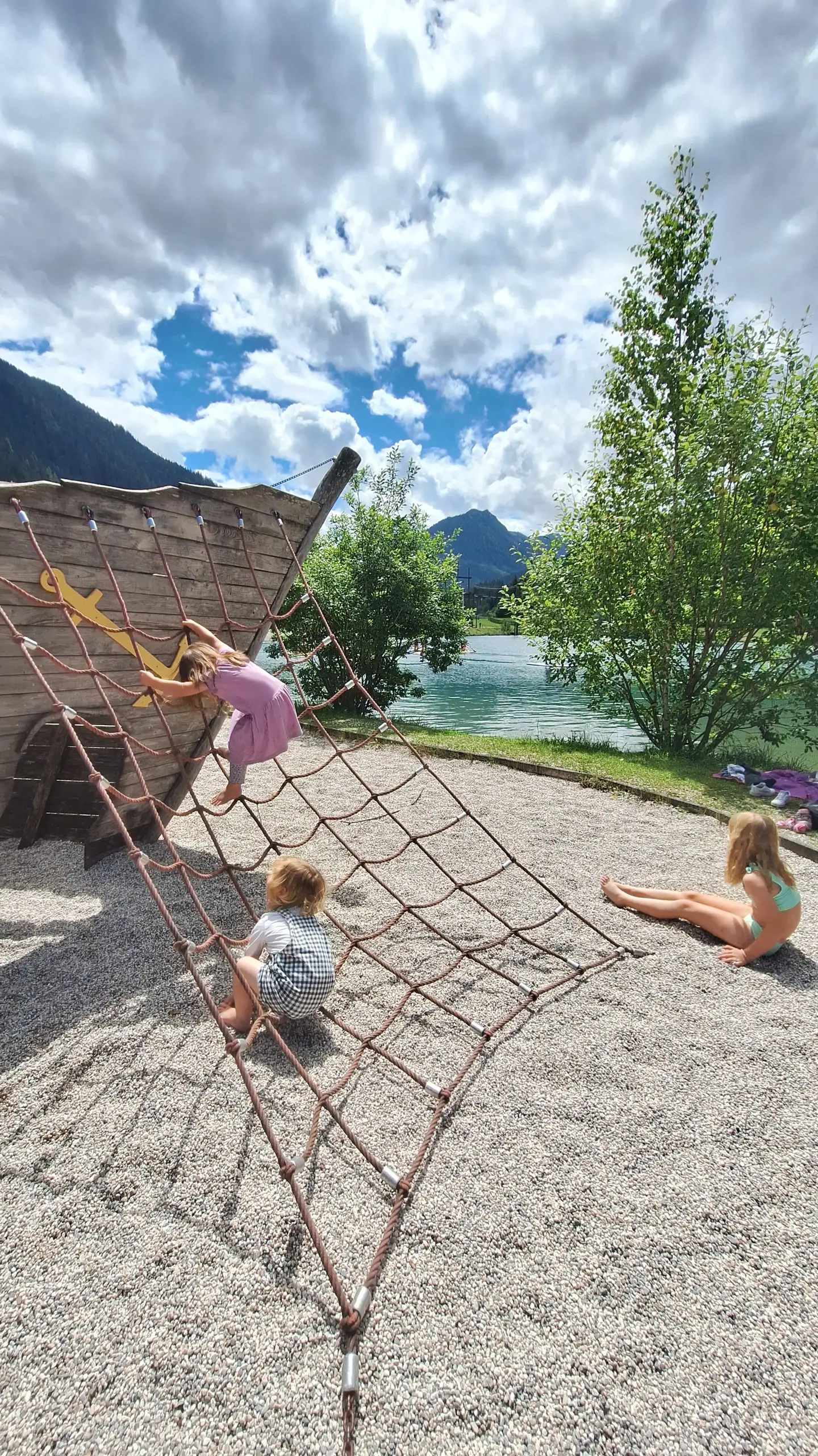 Kinder spielen auf einem Spielplatz am See mit Kletternetz vor Bergkulisse. Sommerliche Aktivität im Freien.