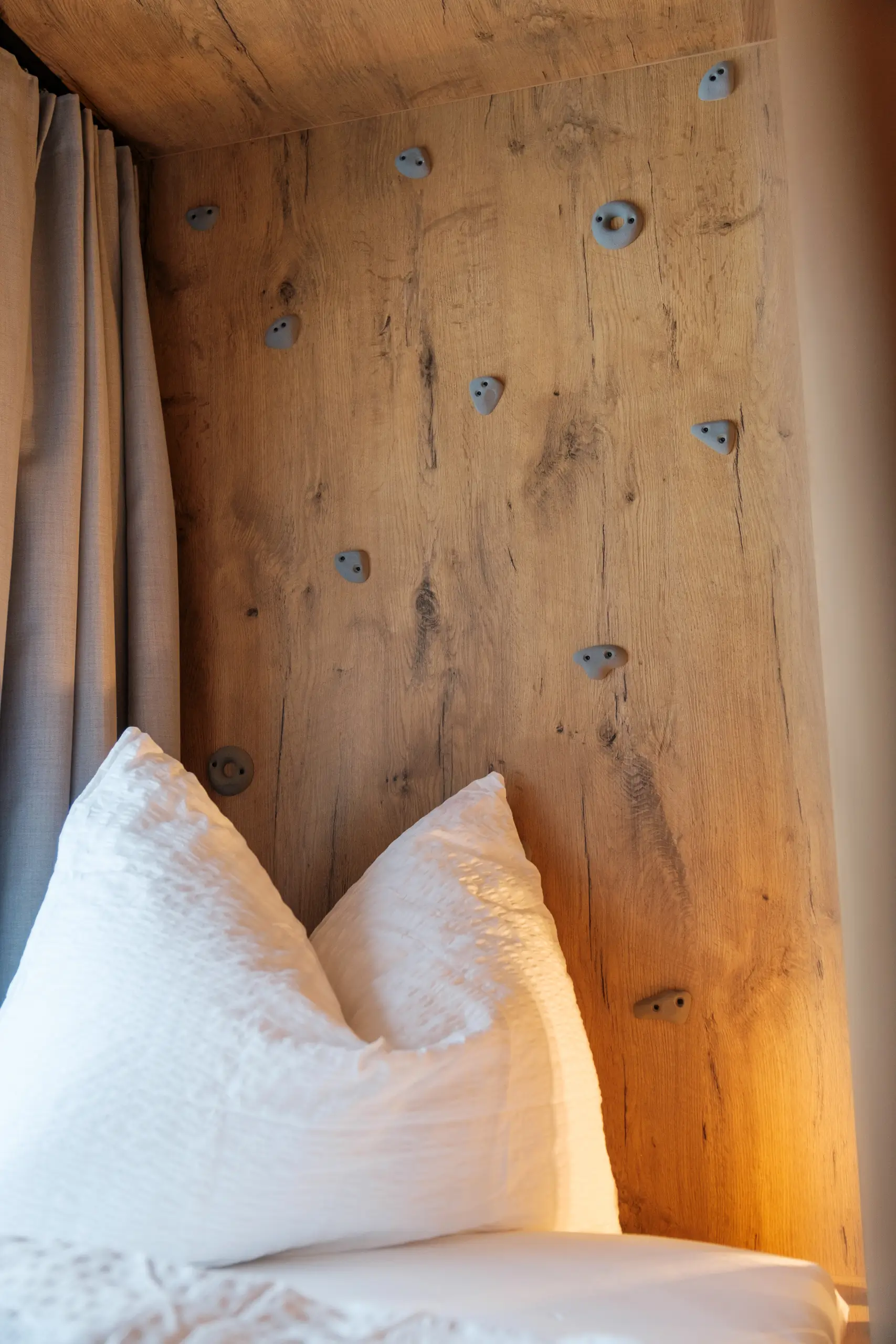 Kletterwand aus Holz mit grauen Griffen über einem Bett mit weißen Kissen.
