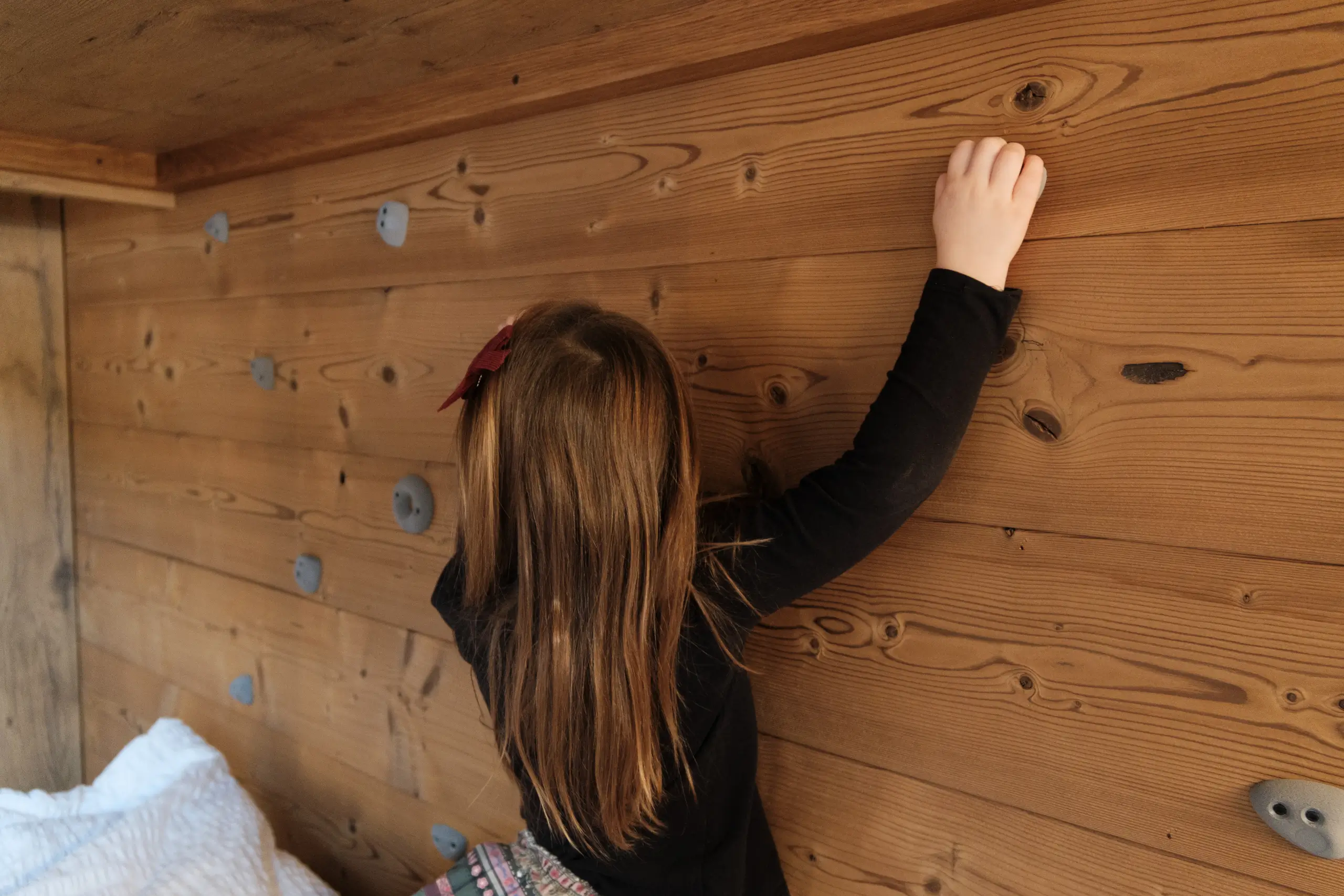 Mädchen klettert an einer Holzwand mit Klettergriffen in einem Innenraum.