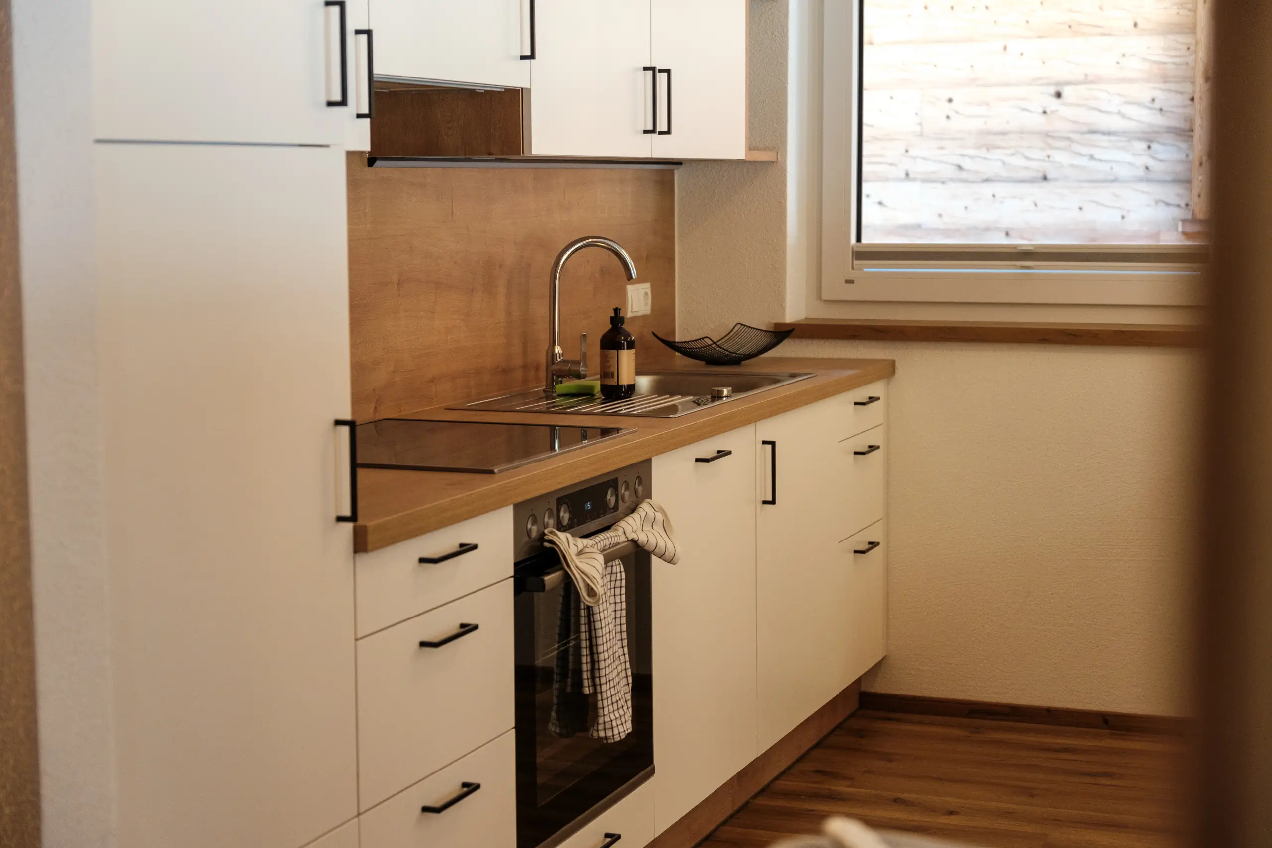 Moderne Küche mit weißen Schränken und hölzerner Arbeitsplatte. Spüle und Fenster.