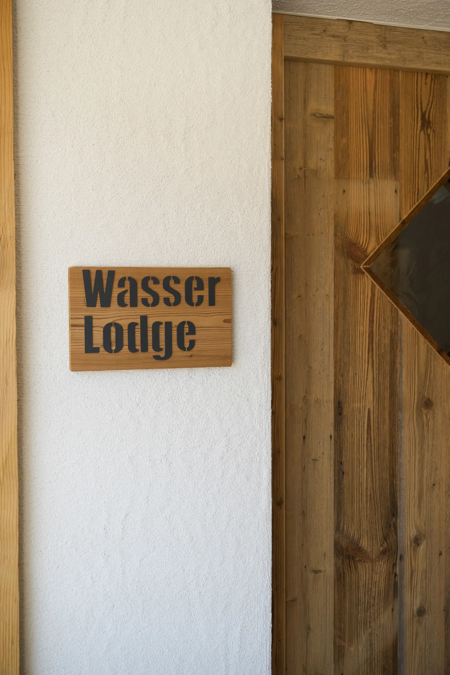 Holzschild "Wasser Lodge" an einer weißen Wand neben einer Holztür.